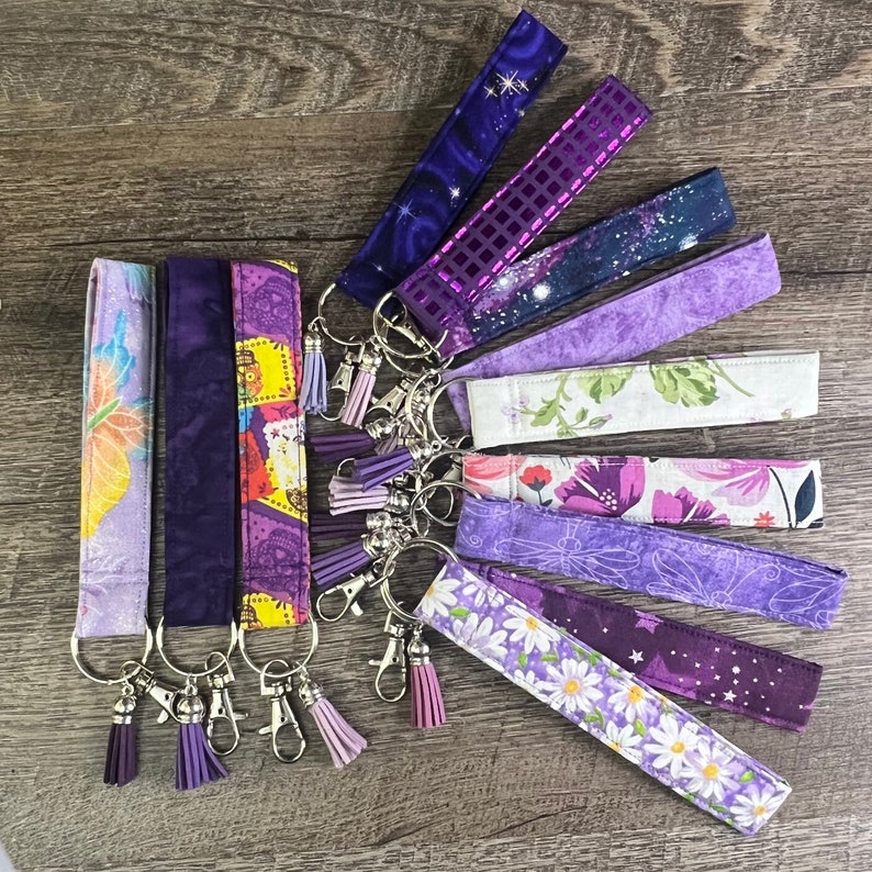 May include: A collection of fabric wrist keychains in various shades of purple, featuring patterns like floral, galaxy, and geometric designs. Each keychain has a metal ring, clasp, and a matching tassel.