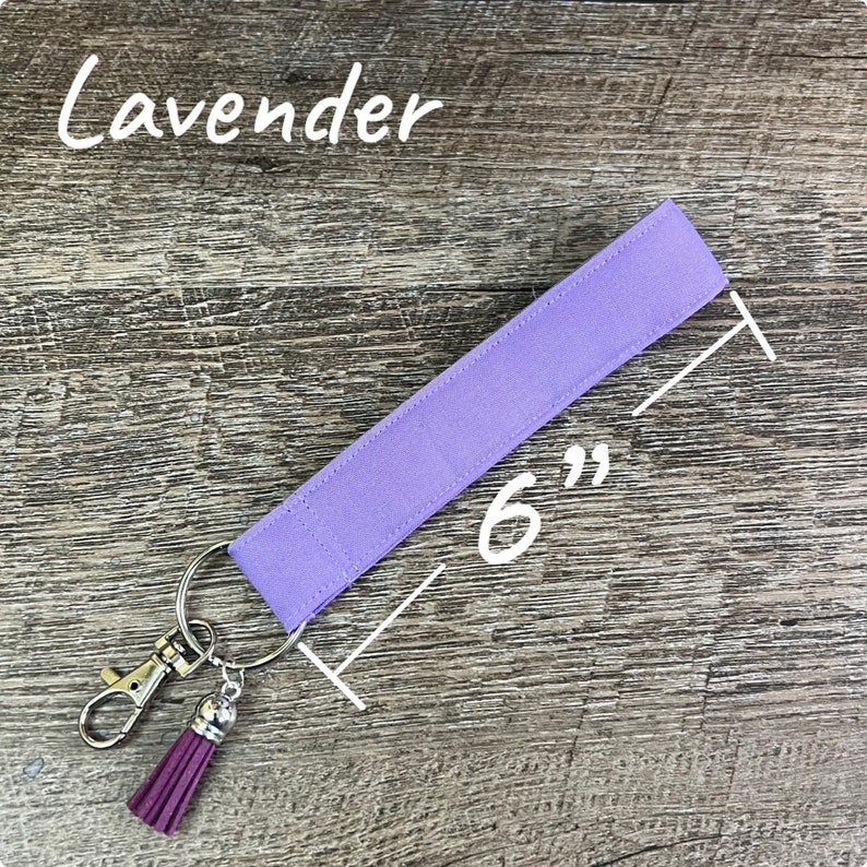 May include: A lavender-colored wristlet keychain, measuring 6 inches long. It features a silver-tone clasp, a key ring, and a matching purple tassel. The word "Lavender" is written in white script at the top.