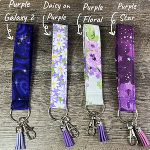 May include: Four fabric lanyards with key rings and tassels. Designs include a dark blue galaxy print, white daisies on purple, purple floral, and a purple star pattern. Each lanyard has a silver-colored key ring and a matching tassel.