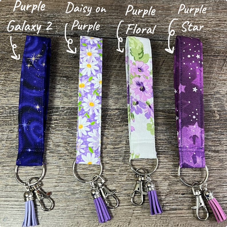 May include: Four fabric lanyards with key rings and tassels. Designs include a dark blue galaxy print, white daisies on purple, purple floral, and a purple star pattern. Each lanyard has a silver-colored key ring and a matching tassel.
