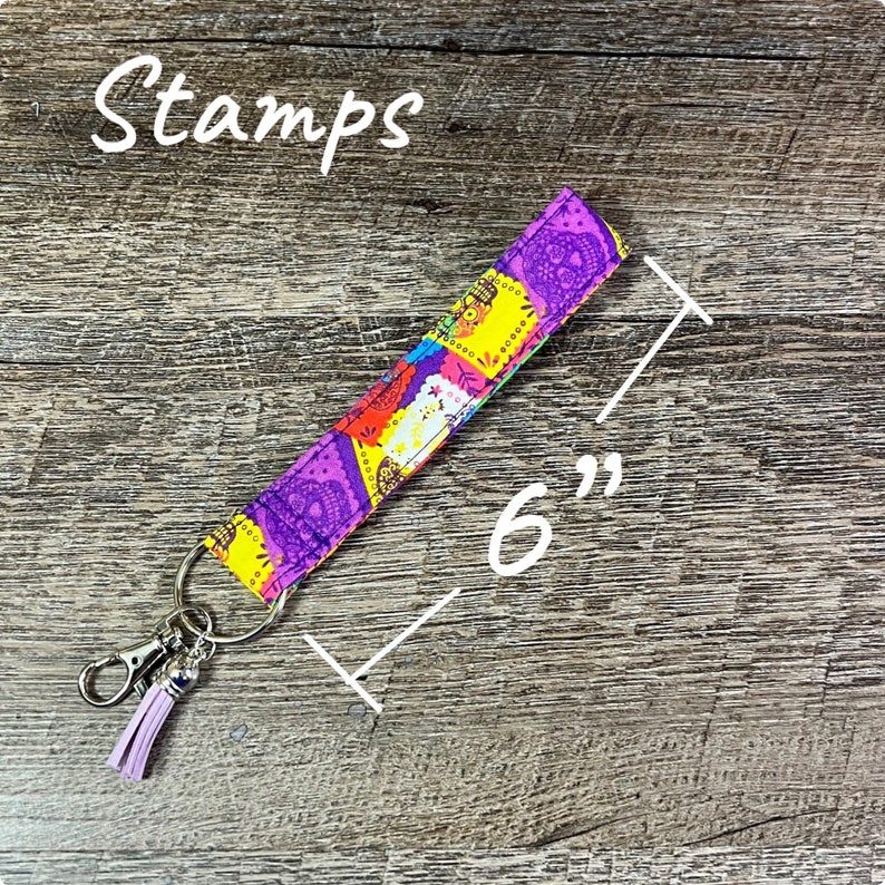 May include: A 6-inch long key fob with a colorful paisley pattern in purple, yellow, red, and blue. The key fob has a silver-colored clasp and a lavender tassel. The word "Stamps" is written in white script.