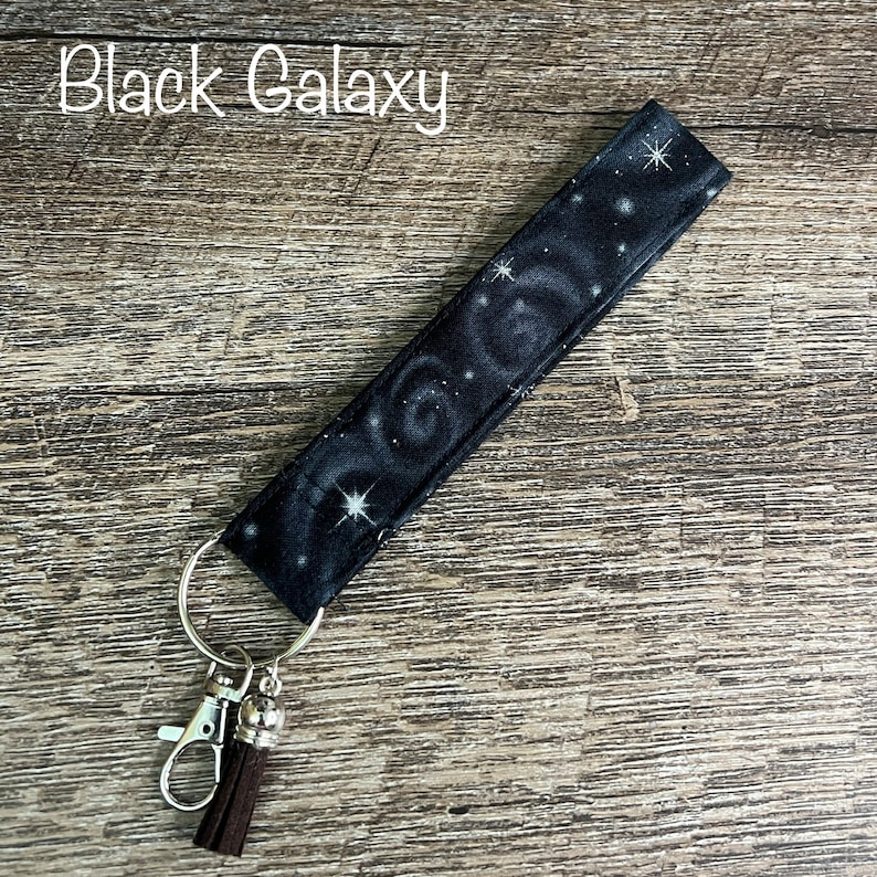 May include: Black galaxy fabric key chain with a silver key ring and a brown tassel. The fabric has a black background with white stars and swirls.
