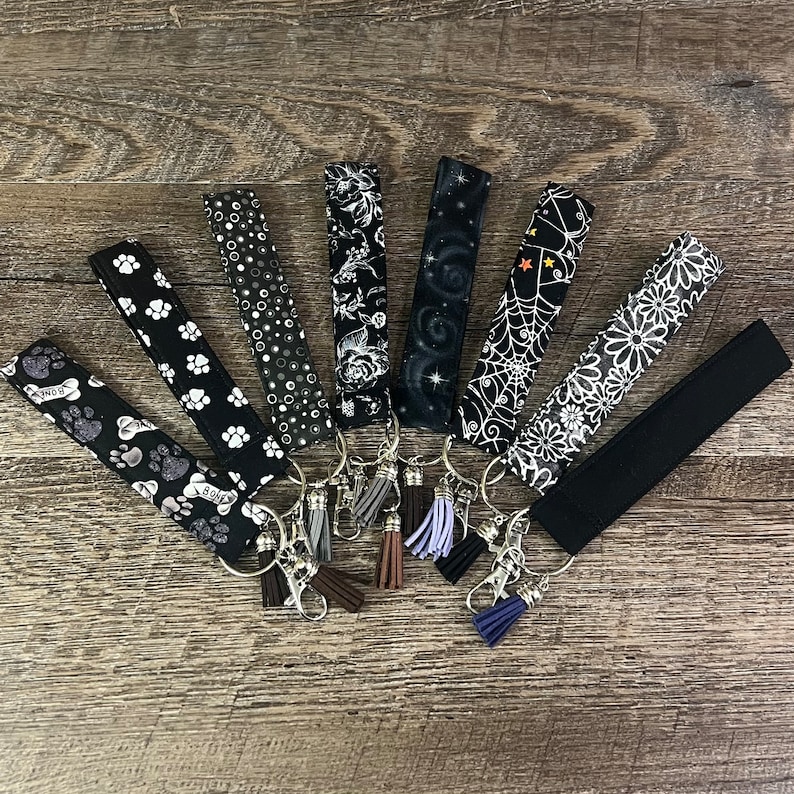 May include: A set of eight black and white keychains with different patterns and designs. Each keychain has a silver key ring and a tassel. The keychains are arranged in a fan shape on a wooden surface.