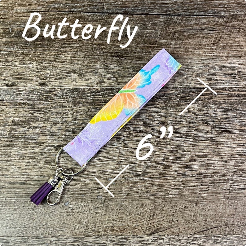 May include: A 6-inch lavender fabric keychain wristlet with a butterfly design in yellow, orange, and blue. The keychain has a silver-colored clasp and a purple tassel.