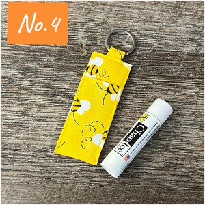 May include: A yellow fabric keychain with a bee pattern and a silver key ring. The keychain is holding a white tube of ChapStick lip balm.