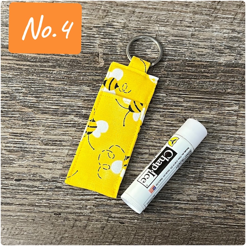 May include: A yellow fabric keychain with a bee pattern and a silver key ring. The keychain is holding a white tube of ChapStick lip balm.