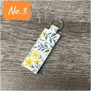 May include: A white fabric key chain with a floral pattern featuring yellow flowers and gray leaves. The key chain has a silver metal ring.
