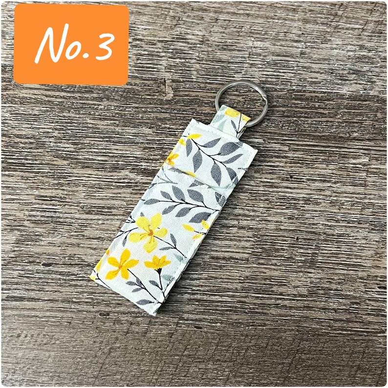 May include: A white fabric key chain with a floral pattern featuring yellow flowers and gray leaves. The key chain has a silver metal ring.