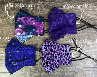 May include: Four face masks with adjustable ear loops are displayed. The masks feature various purple patterns: "Glitter Galaxy", "Indonesian Batik", "Purple Galaxy", and "Purple Cheetah". The masks are made of fabric and are designed to cover the nose and mouth.