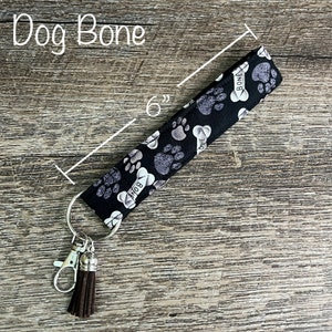May include: Black and white fabric key chain with a paw print and bone design. The key chain is 6 inches long and has a brown tassel.