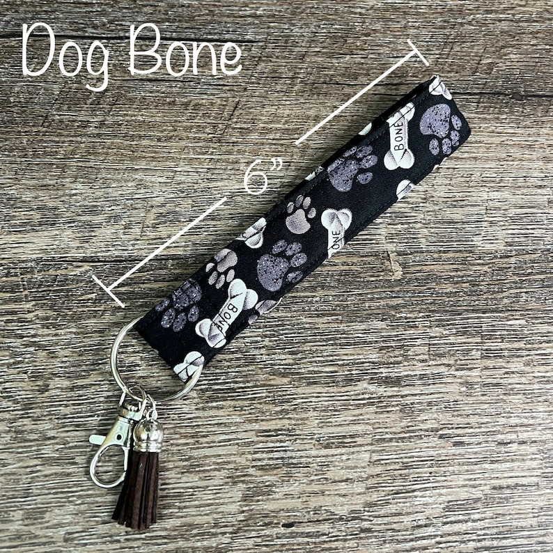 May include: Black and white fabric key chain with a paw print and bone design. The key chain is 6 inches long and has a brown tassel.
