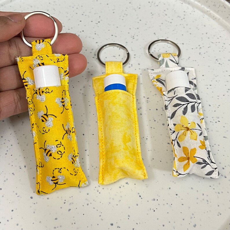 May include: Three yellow and white fabric keychains with silver key rings. Each keychain has a pocket for a lip balm or other small item. The keychains are decorated with different patterns: bees, flowers, and leaves.