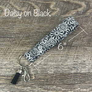 May include: A black and white daisy patterned key chain with a black tassel and a silver key ring. The key chain is 6 inches long. "Daisy on Black" is written in white text on a wooden surface.