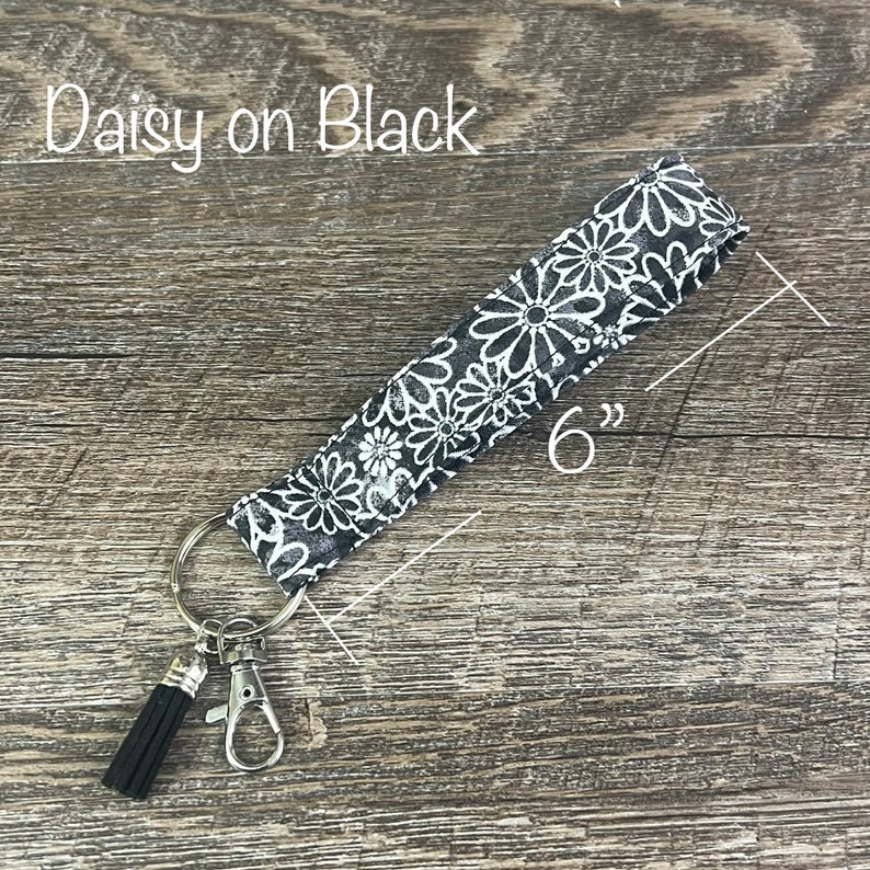 May include: A black and white daisy patterned key chain with a black tassel and a silver key ring. The key chain is 6 inches long. "Daisy on Black" is written in white text on a wooden surface.