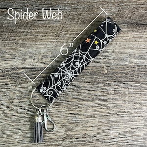 May include: Black and white spiderweb patterned fabric key chain with a silver key ring and a gray tassel. The key chain is 6 inches long.