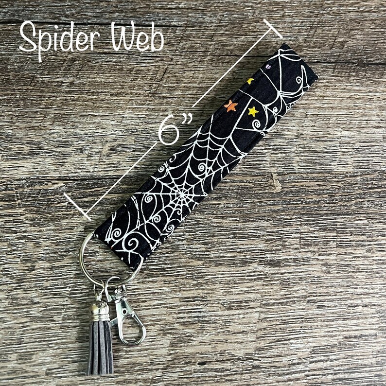 May include: Black and white spiderweb patterned fabric key chain with a silver key ring and a gray tassel. The key chain is 6 inches long.