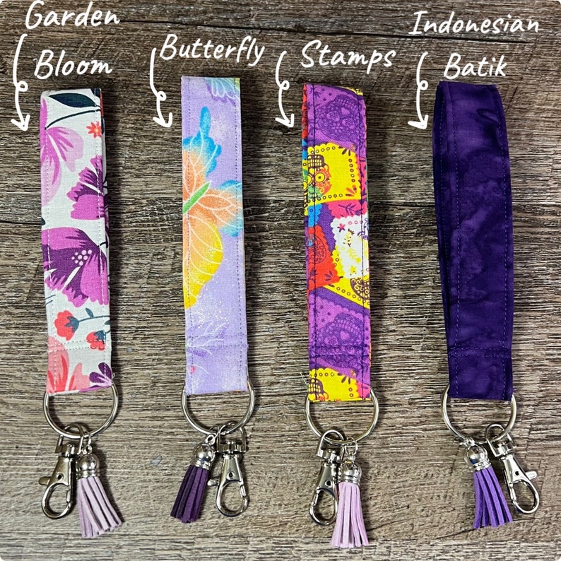 May include: Four fabric wrist lanyards with key rings and tassels. Designs include a floral "Garden Bloom", a butterfly, a colorful "Stamps" pattern, and a purple "Indonesian Batik" print. Each lanyard has a silver-colored clasp.
