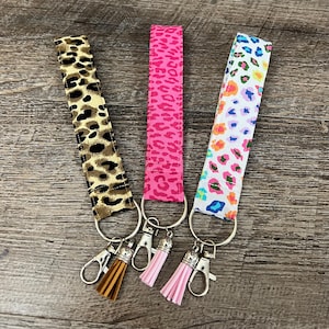May include: Three keychains with different animal print patterns. The keychains are attached to silver key rings with tassels. The patterns are leopard print, pink leopard print, and a multi-colored leopard print.
