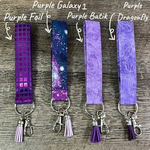 May include: Four keychains with different purple designs. The keychains include a purple foil, a galaxy print, a batik pattern, and a dragonfly pattern. Each keychain has a silver clasp and a matching tassel. The text in the image reads "Purple Galaxy 1", "Purple Foil", "Purple Batik", and "Purple Dragonfly".