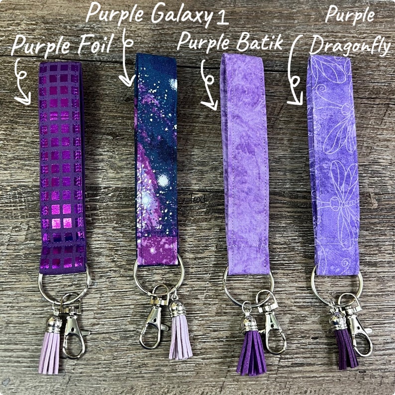 May include: Four keychains with different purple designs. The keychains include a purple foil, a galaxy print, a batik pattern, and a dragonfly pattern. Each keychain has a silver clasp and a matching tassel. The text in the image reads "Purple Galaxy 1", "Purple Foil", "Purple Batik", and "Purple Dragonfly".