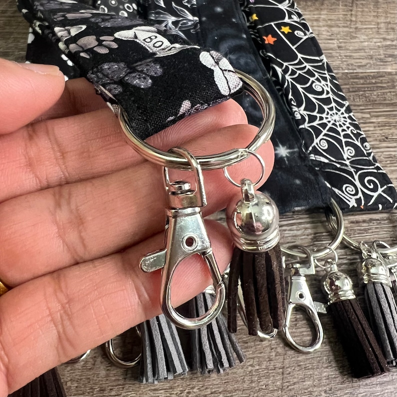 May include: A silver key chain with a silver ring, a silver clasp, and three brown tassels. The key chain is attached to a black and white fabric with a spooky Halloween design.