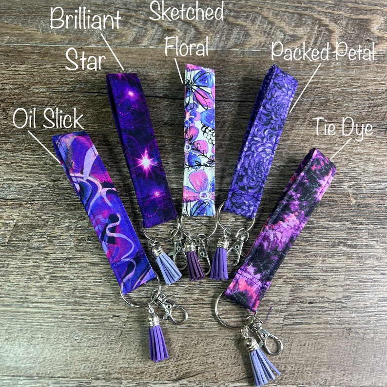 May include: Five fabric keychains with different patterns and colors, including "Oil Slick," "Brilliant Star," "Sketched Floral," "Packed Petal," and "Tie Dye." Each keychain has a silver ring, clasp, and a purple tassel. The background is a wooden surface.