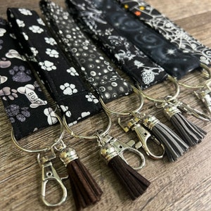 May include: Five black and white patterned keychains with silver key rings and brown and gray tassels. The keychains feature paw prints, polka dots, and a spooky design. One keychain has the word "BOOF" printed on it.