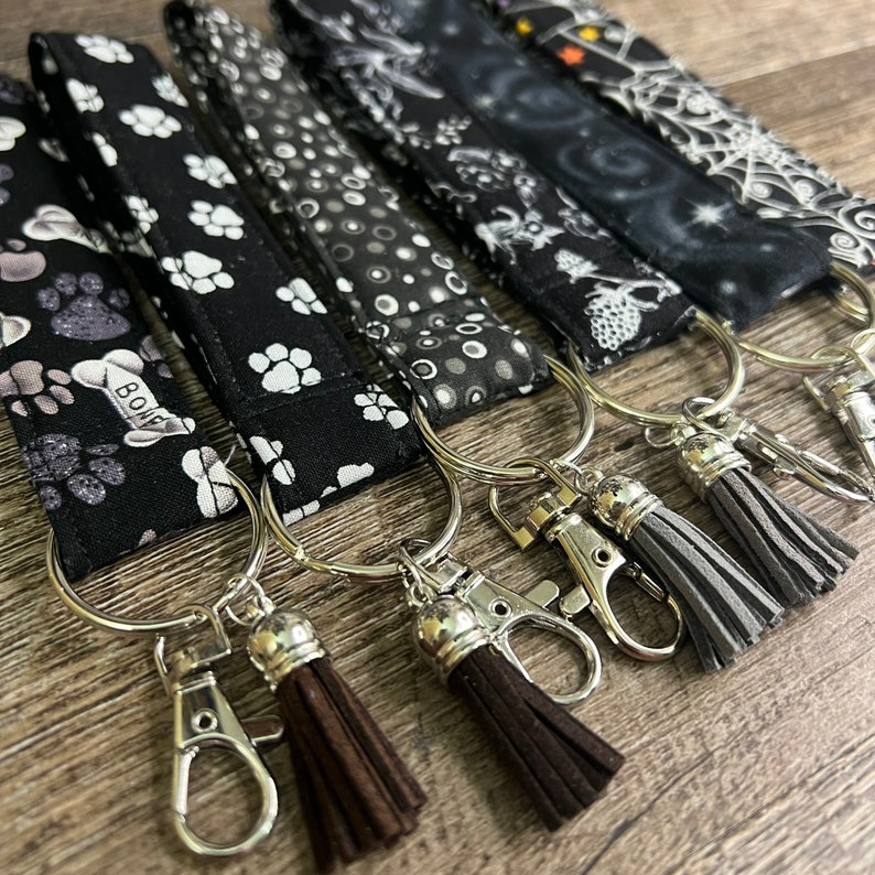 May include: Five black and white patterned keychains with silver key rings and brown and gray tassels. The keychains feature paw prints, polka dots, and a spooky design. One keychain has the word "BOOF" printed on it.