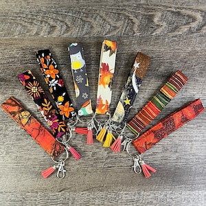 May include: A collection of nine colorful keychains with various autumnal patterns, including owls, leaves, and stars. Each keychain features a metal ring and a tassel.