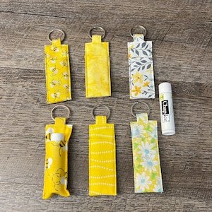 May include: Six yellow fabric keychains with different patterns, including bees, flowers, and polka dots. The keychains are hanging on a wooden surface. A small tube of ChapStick is also visible.
