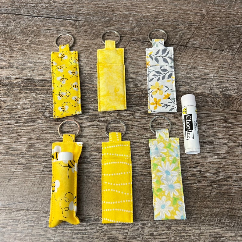May include: Six yellow fabric keychains with different patterns, including bees, flowers, and polka dots. The keychains are hanging on a wooden surface. A small tube of ChapStick is also visible.