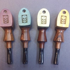 May include: Four wooden pipe tampers with a skull design. Each tamper has a different colored top: purple, teal, cream, and yellow. The stems are dark wood with black tips. The tampers are for pipe smoking.