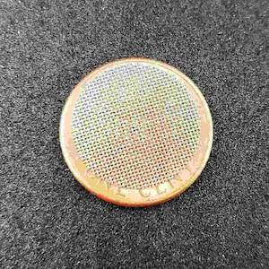 May include: A close-up of a round, copper-colored coin with a mesh pattern in the center. The coin has the word "Liberty" inscribed around the edge.