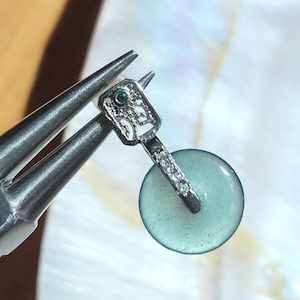 Puede incluir: Un colgante plateado con una parte superior en forma de llave, una pequeña gema verde y una piedra circular azul claro. El colgante está sujeto por unos alicates plateados sobre un fondo blanco iridiscente. El diseño del colgante incluye pequeñas piedras transparentes.