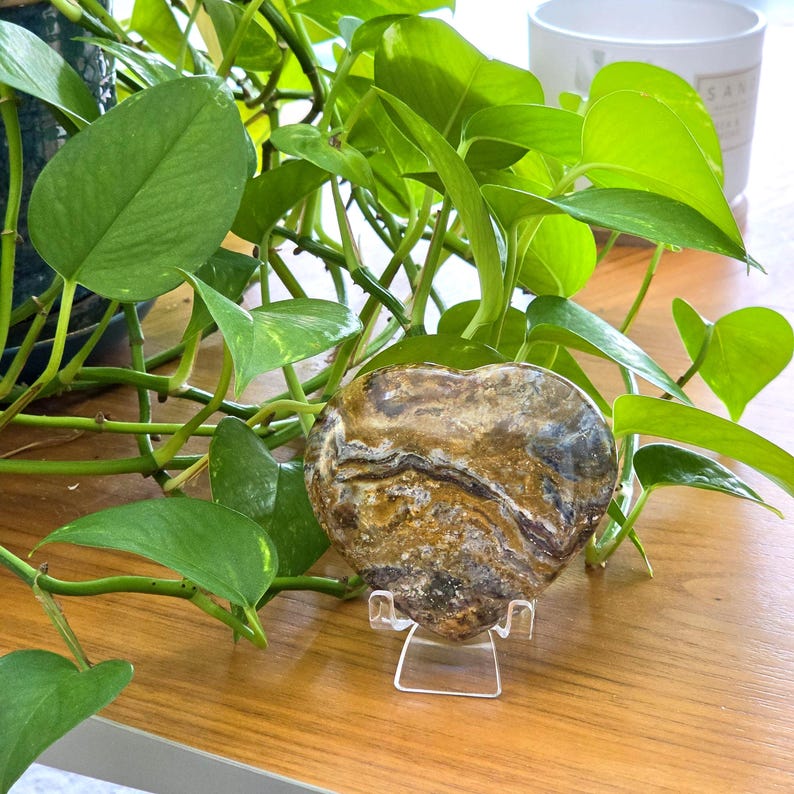 May include: A heart-shaped polished stone featuring swirling patterns of brown, tan, cream, and dark gray. The stone is displayed on a clear acrylic stand, showcasing its smooth, glossy surface and intricate natural mineral banding throughout.