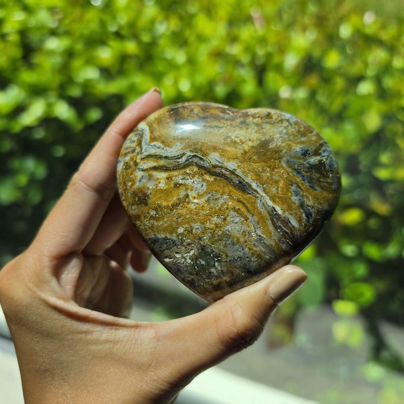 May include: A heart-shaped polished stone featuring a complex pattern of swirling colors including mustard yellow, deep brown, grey, and white. The surface is smooth and reflective, displaying natural mineral veins and organic textures throughout.