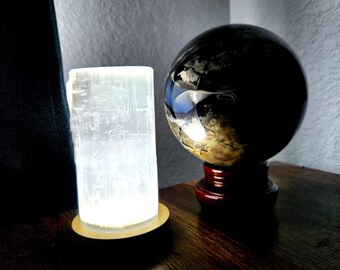 May include: A glowing selenite crystal lamp and a dark, patterned sphere on a wooden surface. The selenite lamp is cylindrical and emits a soft, white light. The sphere sits on a red wooden stand, reflecting light.