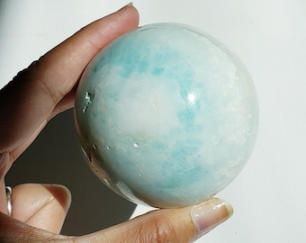 May include: A smooth, light blue and white agate sphere with a polished finish. The sphere is held in a hand.