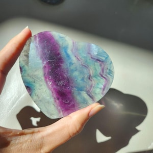 May include: A heart-shaped polished fluorite stone featuring distinct bands of deep purple, translucent light blue, and pale green. The mineral displays a smooth, glossy surface with natural internal colour variations and layered patterns throughout.