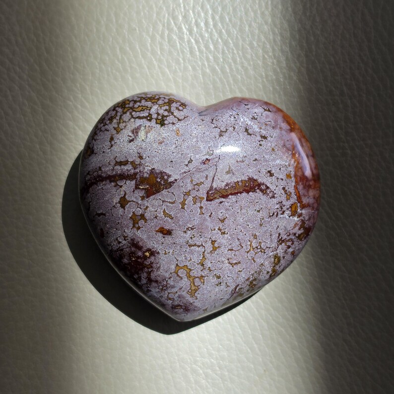 Może przedstawiać: Wypolerowany kamień w kształcie serca z nakrapianym wzorem w odcieniach lawendy, jasnego fioletu i głębokiego brązu. Powierzchnia ukazuje złożoną sieć organicznych, pajęczynowatych inkluzji i żyłek, kt&oacute;re tworzą teksturowany, wielobarwny wygląd.