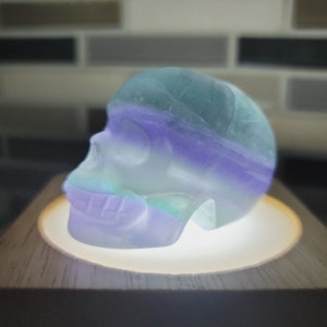 May include: A crystal skull carved from fluorite, with a layered color pattern of green, purple, and white. The skull is displayed on a wooden base with a light shining up from the bottom.