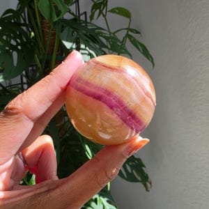 May include: A polished spherical stone featuring translucent layers of warm amber, gold, and deep magenta. The smooth, rounded surface displays natural banding patterns that transition from soft yellow tones to a bold, saturated purple-pink stripe.