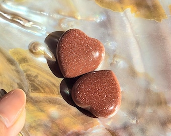 May include: Two heart-shaped, reddish-brown stones with a sparkling, granular texture. The stones are resting on a pearlescent, iridescent surface. The stones are smooth and polished, reflecting light.