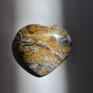 May include: A heart-shaped stone featuring a polished surface with swirling patterns of golden yellow, deep brown, grey, and white. The mineral displays intricate, layered bands and organic textures throughout its smooth, rounded form.