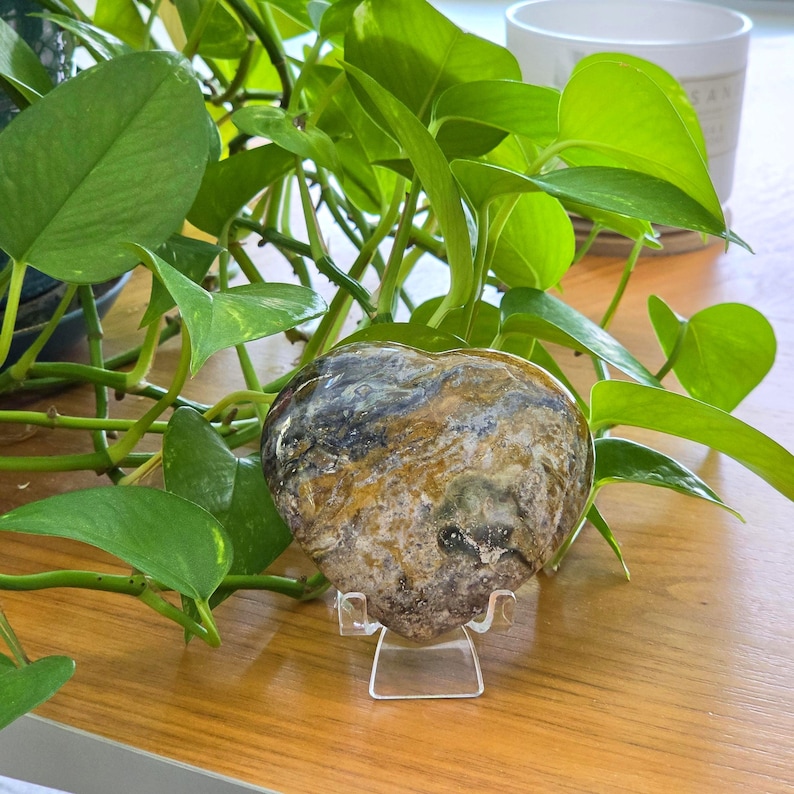 May include: A heart-shaped stone with a polished surface featuring a marbled pattern of brown, tan, gray, and black colors. The stone is displayed upright on a small, clear acrylic stand, showcasing its smooth texture and intricate natural veining.