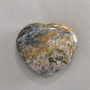May include: A polished heart-shaped stone featuring a complex marbled pattern with swirling colors of ochre, dark gray, white, and muted green. The surface is smooth and glossy, displaying natural mineral inclusions and varied textures throughout.