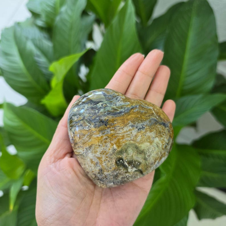 May include: A heart-shaped polished stone featuring a complex pattern of swirling colors including shades of ochre, dark brown, grey, and muted green. The surface is smooth and glossy, displaying natural mineral veins and organic textures throughout.