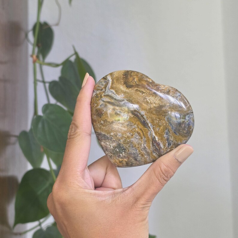 May include: A polished heart-shaped stone featuring a complex marbled pattern with swirling colors of golden brown, deep ochre, dark charcoal, and hints of muted blue. The surface is smooth and reflective, showcasing natural mineral veins throughout.