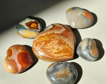 May include: Five polished heart-shaped stones of varying sizes arranged in a cluster. The stones feature natural patterns in shades of orange, brown, grey, and cream. The largest heart is centrally located, surrounded by four smaller heart-shaped stones.