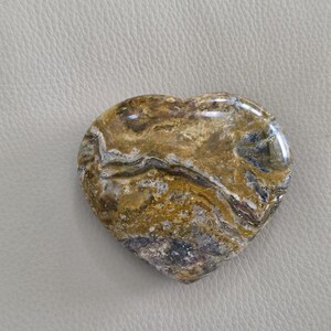 May include: A heart-shaped polished stone featuring a complex pattern of swirling colors including shades of golden brown, dark gray, and white. The surface is smooth and reflective, showcasing natural mineral veins and organic textures throughout.